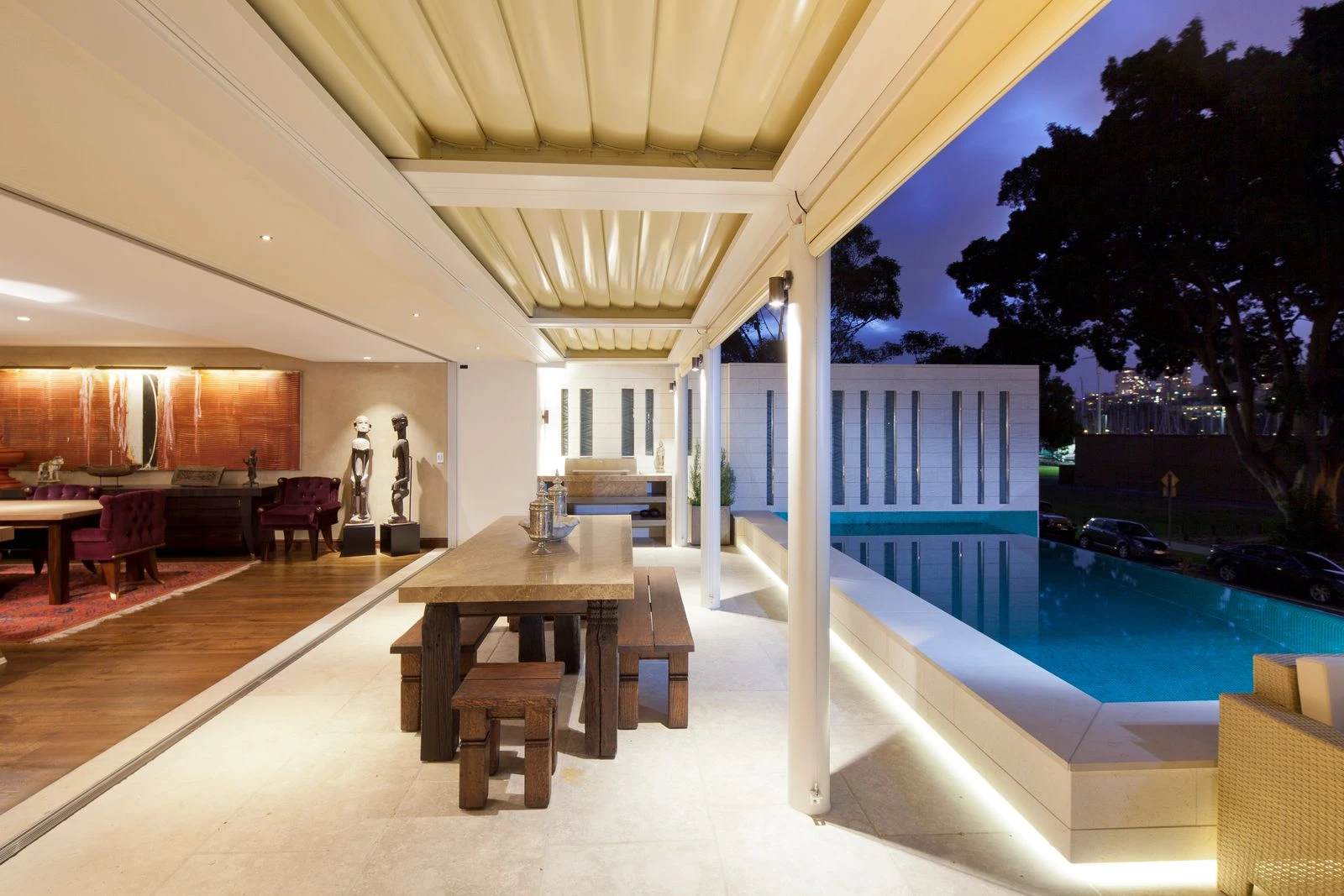 Covered alfresco dining room beside a pool at dusk, lit from the house behind